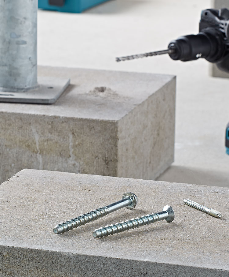 Produktbild von zwei Betonschrauben auf einem Betonsockel im Hintergund eine Bohrmaschine